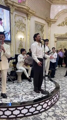 A group of musicians is performing on a stage in a room with ornate decor. The room has golden accents on the walls and ceiling, and a large screen is displaying an image of a man playing a guitar. The musicians are all dressed in white or light-colored clothing. A man in the foreground is singing into a microphone, while another man to his left plays a clarinet. In the background, a man is playing a drum, and others are playing guitars and keyboards. The floor is covered with a patterned carpet. There are audience members visible in the background on the right side of the frame.