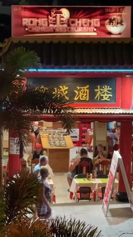 The video shows the exterior of a Chinese restaurant called Rong Cheng Chinese Restaurant at night. The restaurant has a red and gold facade with neon lighting. There are several tables and chairs set up outside, and people are seated at some of them. Some people are walking past the restaurant. The overall atmosphere appears to be casual and lively.