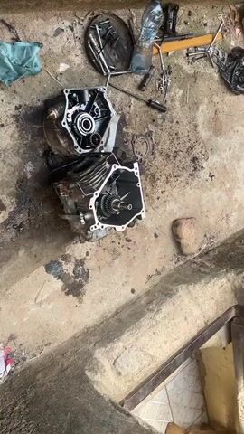 The video shows a disassembled engine on a concrete floor. The engine is open, exposing gears and internal components.  Tools, a water bottle, and a gasket are scattered around the engine parts. The floor is stained with oil.  There is no person visible. The perspective is from directly overhead, with a slight rotation.  A tiled surface and a metal object are seen partially visible at the bottom right of the frame.