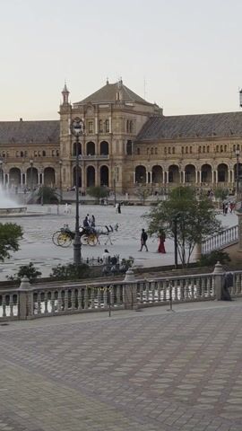The video shows a wide view of the Plaza de España in Seville, Spain. In the foreground is a brick-paved walkway with a low stone railing. Behind the railing is a canal or small body of water, and beyond that is the Plaza itself. The Plaza is a large open space with a light-colored paved surface. A large fountain is prominently featured in the middle of the Plaza. 

The Plaza is bordered by a grand, semi-circular building with arches, columns, and towers. People are seen walking and moving around the Plaza. A horse-drawn carriage is visible, moving through the Plaza. One individual dressed in a long red dress walks across the Plaza. 

The sky is overcast, suggesting the time of day is either early morning or late afternoon. The overall impression is of a spacious and architecturally rich public square, bustling with activity.