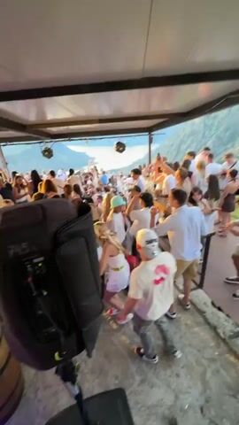 This video shows a lively party atmosphere with many people dancing and socializing under a covered outdoor area. The camera pans across the scene, capturing glimpses of individuals dressed in casual attire, some wearing masks. There are speakers and barrels visible, suggesting a DJ setup and bar service. The background reveals a scenic view of hills or mountains, illuminated by natural light. The overall impression is one of a vibrant celebration.