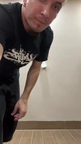 A young man with light skin and dark hair is dancing in a room. He is wearing a black t-shirt with a white graphic design and dark shorts. He has on white sneakers. The room has a light-colored wall and a wooden floor. The man is in the center of the frame, moving his body with energy and expression. His face shows focus and engagement with the activity. The lighting is bright, suggesting it is daytime or a well-lit indoor space. The scene appears to be a casual setting, possibly a home or a dance studio. There is a red tag on the sleeve of the man's t-shirt.