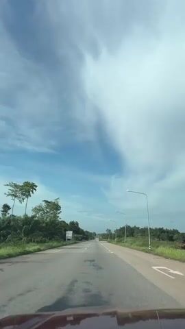 The video shows a view from the perspective of someone inside a moving vehicle, looking forward down a paved road. The road appears to be a highway or a significant rural road with two lanes in each direction, separated by a median. White lane markers are visible on the road surface. The road is lined with trees and vegetation on both sides. The sky is blue with scattered white clouds, some of which are large and wispy. Streetlights are visible at intervals along the side of the road. There is a sign visible on the left side of the road. The hood of the vehicle the footage is being filmed from is visible at the bottom of the frame, reflecting the sky. The overall environment is sunny and appears to be outdoors.