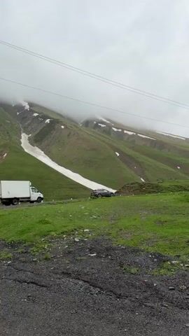 A truck and a silver sedan are on a winding mountain road. The truck is white and parked on the right side of the road. The sedan is driving on the left side of the road. The road is paved with gravel and is surrounded by green grass. In the background, there are steep, green mountains with patches of snow. The sky is overcast with clouds. The video appears to be filmed from a moving vehicle.