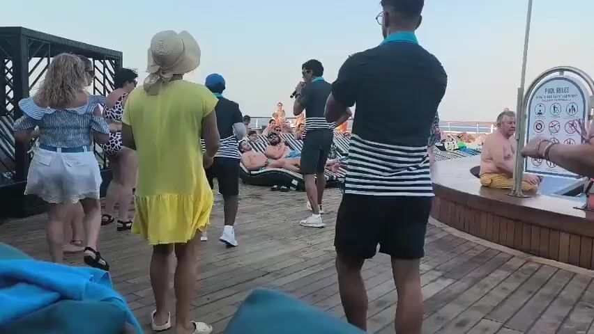 The video shows a group of people dancing on a wooden deck. The sky is overcast and appears to be late in the day. In the foreground, a woman in a yellow dress is dancing, her dress is twirling around her. To her left, a woman in a white top and denim shorts is also dancing. In the center of the frame, two men wearing black shirts with striped accents and black shorts are leading the dance. The man on the right is wearing glasses. To the left of them, another person in a black shirt with stripes is playing an instrument. Several other people are sitting on lounge chairs in the background, some are in swimsuits. To the right, a woman in a patterned swimsuit is dancing. Further in the background, there is a swimming pool with people in it. A sign with pictograms is visible near the pool. The overall atmosphere seems to be a fun, outdoor event.