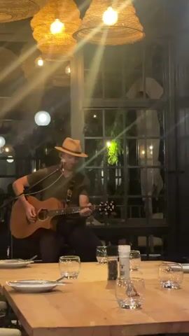 A musician is playing guitar and singing in a restaurant, sitting on a table set for a meal. He is wearing a hat and olive green shirt.  Two large straw light fixtures hang above the table, along with another light in the background.  The table is wooden and surrounded by clear glasses, plates, and a pepper grinder.  Behind the musician is a large window with dark trim looking out onto a dark background with some greenery.
