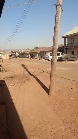 The video shows a street scene in a somewhat rural or developing area. The street is primarily dirt with some patches of asphalt. There's a wooden utility pole in the foreground, casting a long shadow across the street. Overhead, there are power lines stretching across the sky. A person is walking down the street in the distance. Several buildings line the street, including a two-story structure with a balcony. There are a few vehicles parked along the street, including a white van. The sky is clear and blue. The overall setting appears to be sunny and dusty.