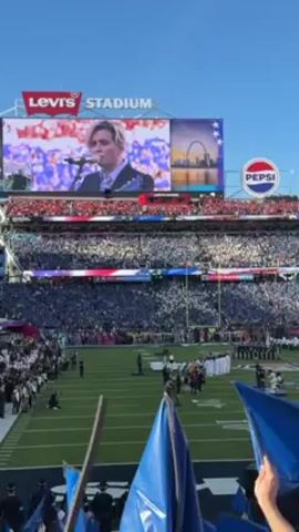 The video shows a football stadium filled with people, with a large screen showing a person singing and playing guitar. The stadium is Levi's Stadium, as indicated by the sign. There is also a Pepsi logo visible. Below the screen, on the field, there are people standing in formation, some wearing white robes, and others in what appear to be military uniforms. In the foreground, there are blue flags being held. The overall atmosphere suggests a ceremony or performance before a sporting event. The sky is clear and blue.