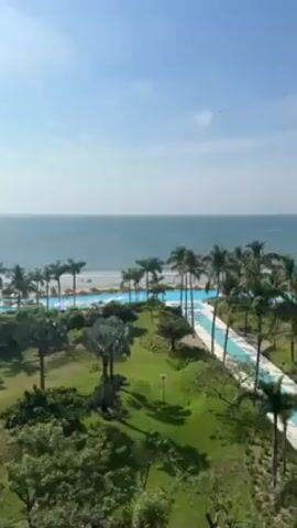 The video shows a scenic view of a beachside resort under a clear, partly cloudy sky. A large turquoise pool snakes through lush green grass and palm trees. Beyond the pool, the sandy beach meets the ocean, where a few people can be seen. The camera remains still.