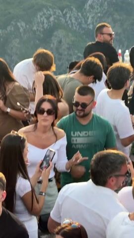 The video shows a gathering of young adults outdoors, possibly on a terrace or a hilltop, with a mountainous landscape visible in the background. The lighting suggests it is daytime, with some atmospheric haze. People are dressed casually, many in sunglasses, and seem to be socializing. Some individuals are holding drinks and phones. There's a sense of a party or a social event, with people interacting, talking, and looking around. The camera is positioned slightly above and behind the crowd, capturing a lively group dynamic.