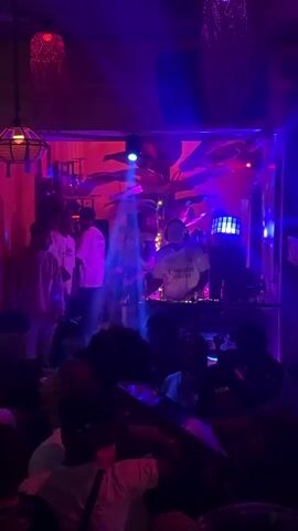 The video captures an indoor party or club scene at night in Shenyang City, China. The setting is dimly lit but vibrant, with dynamic red, blue, and green disco lights casting colorful streaks and patterns across the room and onto the crowd. Several decorative hanging lamps are visible, adding to the ambiance.

The primary focus is a DJ positioned behind a set of turntables and a mixer, actively playing music. The DJ is wearing a white T-shirt with "Emirates Fly Better" printed on it. Surrounding the DJ and filling the space are numerous people, mostly seen from behind or in silhouette due to the low lighting. They appear to be dancing, socializing, and enjoying the music, with some holding drinks. Large green plants are strategically placed, enhancing the club's tropical or lounge aesthetic. The overall atmosphere is energetic and lively.