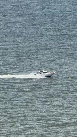 The video depicts a white speedboat moving rapidly across a body of water, leaving a white wake behind it. The water is a grey-blue color and slightly choppy. In the background, a landscape of rolling hills and snow-capped mountains is visible beneath a cloudy sky. The camera pans upward to reveal more of the mountain range and sky. The boat maintains a steady course across the water throughout the video.
