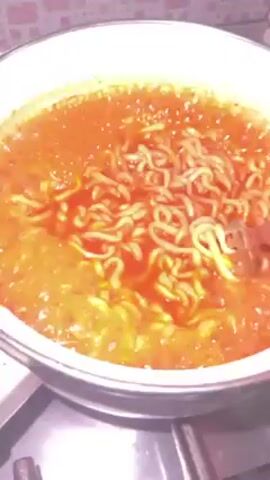 A high-angle shot shows a white pot with a silver rim sitting on a black stovetop. The pot is filled with a reddish-orange liquid, likely a broth, containing noodles. A hand, presumably belonging to a person standing behind the camera, is seen using a metal fork to stir the noodles within the liquid. The noodles appear to be instant ramen-style, with a wavy texture. The background features a kitchen backsplash composed of small, light-colored tiles.