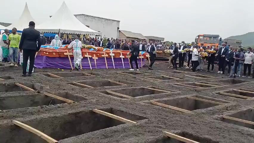 The video depicts a funeral service with a row of caskets laid out in front of multiple dug graves. People are standing around, some wearing suits and others wearing protective suits. Some are carrying flowers.  There are two large white tents and a truck in the background. The sky is overcast.