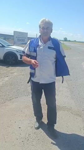 A man stands on a dirt road, wearing a blue vest over a white shirt and dark pants. A white vehicle is parked behind him to the left. He smiles and gestures with his hands. The video then cuts to a different man, wearing a green vest over a black shirt and black pants, standing with his hands on his hips. A white SUV is parked behind him to the right. Another dark-colored vehicle is parked behind him to the left. The scene appears to be an outdoor setting with a fence in the background.