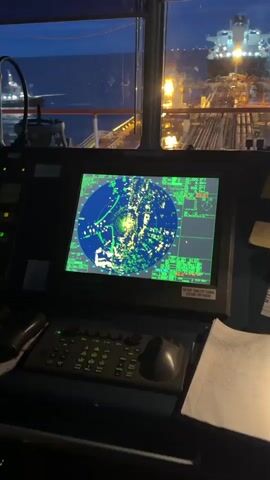 The video shows the interior of a ship's bridge at night. A large radar screen displays numerous green and yellow dots, indicating other vessels and navigational markers. Outside the windows, the dark sea is visible, along with the lights of another large ship, possibly a tanker. The scene suggests the ship is at sea, possibly near other vessels, and navigating through darkness. The camera is positioned looking out of the bridge windows, with the radar console in the foreground.
