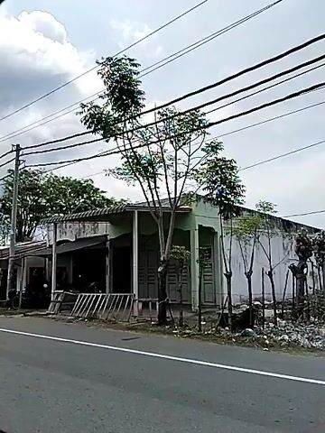 The video captures a static view from a moving vehicle traveling along a paved road. On the right side of the road, there are several roadside structures, including what appears to be a small shop with a corrugated metal roof and a stall. A person is riding a motorcycle, wearing a helmet and a patterned shirt, moving away from the camera. Overhead, multiple power lines are strung between utility poles. The environment is somewhat rural or semi-urban, with trees, vegetation, and scattered debris visible along the roadside. The sky is cloudy, suggesting overcast weather. In the background, further structures and trees are present. The video continues to show the roadside scenery as the vehicle moves.
