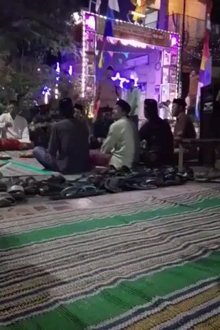 The video shows a group of people sitting on mats in what appears to be an outdoor setting at night. There are several pairs of footwear, including sandals and flip-flops, scattered in the foreground. In the background, there is a decorated archway with lights and banners, possibly indicating a celebration or event. Several individuals are standing or sitting near the archway, and some appear to be speaking or performing. The overall atmosphere suggests a community gathering or religious event.