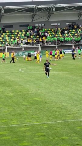 A football match is nearing its end. A player in a black and white uniform stands on the green grass of the field, holding a football. In the background, spectators are seated in the stands, which are a mix of yellow and green. Some people are gathered near the sideline. A large screen displays the score, indicating the match is at 90 minutes plus 4 minutes of added time, with the score being 1-0. Several individuals, possibly staff or media, are positioned around the field and near the screen. The environment is a football stadium with a well-maintained grass pitch and tiered seating.