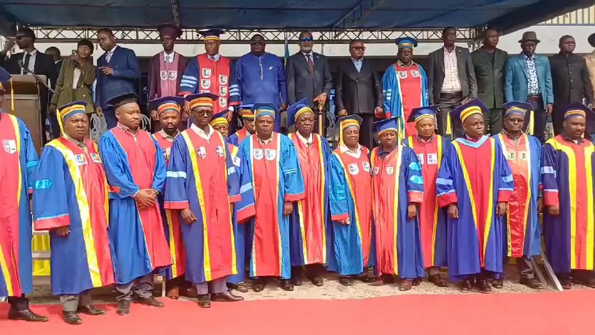 A wide shot shows a group of around 25 people standing on what appears to be a stage. The people are dressed in academic gowns of blue, red, and yellow. Some wear hats that match the gowns. Behind them, a smaller group stands on a higher level of the stage, dressed in suits and more academic gowns. A blue awning covers the stage. A small, bright object hangs from the awning. The people in the academic gowns are arranged in rows and are looking at the camera. Some of the people behind them also look towards the camera. The environment appears to be outdoors, possibly a graduation or academic ceremony.