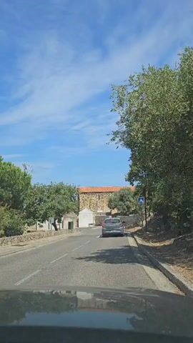 The video shows a view from inside a vehicle traveling on a road. A silver car is in front, driving away from the camera. The road is paved and appears to be in a rural or semi-rural area, with trees and some stone walls lining the sides. In the background, there is a building with a red tile roof and stone walls, possibly a church or an old stone structure. There are white buildings and a white bench visible on the left side of the road. The sky is blue with scattered white clouds. A pedestrian crossing sign is visible on the right side of the road. The road curves slightly to the left.