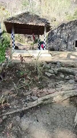The video shows an outdoor scene. In the foreground, there is rocky ground with some dry leaves and branches. In the middle ground, there is a small, open-sided structure with a thatched roof, supported by checkered pillars. Inside the structure, two individuals are seated. To the right of the structure is a large, rough-hewn rock face with a rectangular opening. The background consists of a rocky hillside with sparse vegetation. The lighting suggests it is daytime, with sunlight casting shadows.