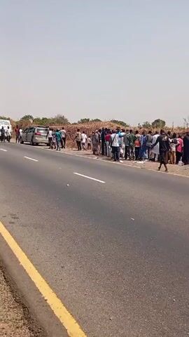The video shows a rural road with several vehicles parked along the shoulder, facing the same direction. A group of people stands near the roadside, some on an embankment overlooking the road. The ground around them is littered with white paper. The overall scene suggests the aftermath of an event, possibly an accident or incident, that has drawn a crowd.
