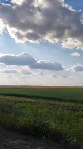 The video displays a vast, open landscape under a dynamic sky filled with white, puffy clouds casting shadows and allowing shafts of sunlight to break through. In the foreground, there is a dirt or gravel road on the left side of the frame. To the right of the road, tall green grass and some wildflowers are visible, swaying slightly. Beyond the immediate foreground, there are fields of green crops and then a distant field of brown or dry vegetation, stretching to the horizon. The overall impression is of a rural or agricultural setting on a partly cloudy day.