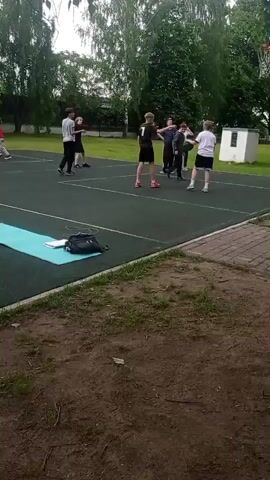 The scene is a video taken on an outdoor basketball court. The camera is static and positioned just off the court, facing the action. There are five or six teenagers on the court. One boy dribbles a basketball, while others are standing around.  A light blue yoga mat and a black bag are lying on the court near the bottom left of the frame. The court has green flooring with white lines marking the boundaries. Behind the court, there is a grassy area and trees. A white square building is visible at the far end of the court. The sky is overcast.