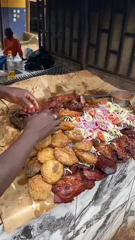 This media captures a vibrant outdoor food stall in Barakin, Nigeria. The primary subjects are an abundance of prepared food, presented on large sheets of brown paper. Prominently featured are piles of golden-brown fried fritters, alongside various cuts of grilled or roasted meats that appear dark and savory. A fresh salad of shredded cabbage, sliced tomatoes, and red onions is artfully placed among the meats. A small metal bowl containing a bright orange sauce, possibly a condiment or seasoning, sits nearby, along with a plastic container.

In the background, two people are visible. One, a young boy in an orange hoodie and blue pants, sits on a concrete block, facing away from the camera. Another person, partially obscured, is seen in the distance, wearing a red jacket. To the left, a motorcycle is parked, suggesting the everyday bustle of the location. The scene is lit by natural daylight, indicating it is likely daytime, though the specific time is not clear. The setting appears to be an informal, possibly roadside, establishment, with a tiled surface in the foreground and a fence or structure with wooden slats in the background. The overall impression is one of a lively food service environment, showcasing local culinary offerings. No visible text is present in the media.
