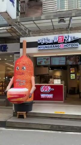 The camera pans slowly from left to right across the street, showing storefronts in front of which there are promotional objects. On the left, the first storefront is for Myungrang Hotdog. There is a smiling corn dog figure in front of the store. Next to it, the adjacent storefront advertises Fried Chicken Master.