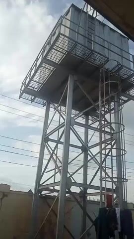 This is a still image, likely captured from a video, showing a tall, metal water tower. The tower is supported by a complex lattice of metal beams, creating a sturdy and industrial aesthetic. A ladder with a protective cage ascends the side of the tower, suggesting it is meant for maintenance or access. Atop the tower sits a large, rectangular water tank, also constructed from metal. A grated platform surrounds the base of the tank, providing a walkway.

In the foreground, clothes are hanging on a line, suggesting a domestic setting or an area where laundry is being dried. The clothes appear to be a mix of dark and lighter colors, and they are casually draped, indicating a relaxed atmosphere.

The background reveals a cloudy sky, hinting at a potentially overcast or breezy day. The presence of power lines crisscrossing the sky further reinforces an outdoor, possibly urban or semi-urban, setting. In the distance, a green tree is visible, adding a touch of nature to the scene. The overall impression is one of functionality and everyday life, with the imposing water tower serving as a prominent landmark against the backdrop of the sky and surrounding environment. There is no visible text in the image.