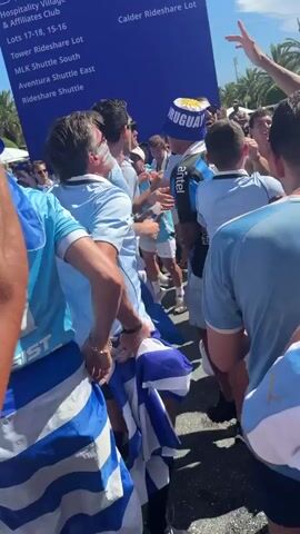 A crowd of people, primarily men, are gathered outdoors, appearing to be exiting a stadium or event. Many are wearing light blue jerseys and hats, some with the word "URUGUAY" visible, suggesting they are fans of the Uruguayan national team. They are holding a large blue sign that reads "Stadium Fan Experience" and lists various locations and services, including "Hospitality Village & Affiliates Club," "Lots 17-18, 15-16," "Tower Rideshare Lot," "MLK Shuttle South," "Aventura Shuttle East," "Rideshare Shuttle," and "Ticket Resolution Point," with "Lots 6, 10-11" and "Calder Rideshare Lot" also listed. Some individuals are raising their arms in celebration or to capture photos with their phones. The atmosphere seems energetic and festive, with clear blue skies and sunlight indicating it is daytime, likely warm weather. Palm trees are visible in the background, and the ground appears to be asphalt, suggesting an outdoor parking lot or pathway. The overall scene depicts a group of enthusiastic fans after a sporting event or concert.