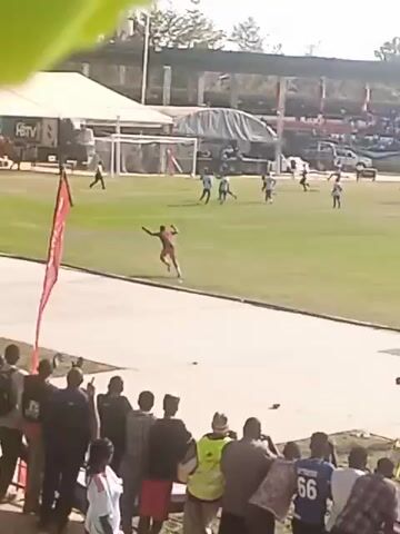The video shows a soccer match in progress. A player in a dark jersey is airborne near the goal. Other players in green and white jerseys are visible on the field. A crowd is watching from the stands, and a person in an orange vest is running along the sidelines. There are tents and vehicles visible in the background beyond the field. The scene is captured from the perspective of someone in the stands.