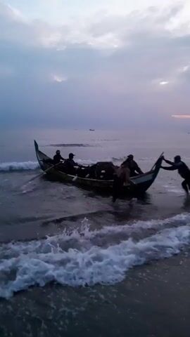 The video shows a small fishing boat with several people in it being pushed into the water from a beach. Two people are actively pushing the boat, while the others are already aboard. The boat has a long oar extending from its side. The ocean is choppy with waves, and the sky is overcast with a hint of light on the horizon. The overall atmosphere is one of early morning or late evening as fishermen embark on their task.