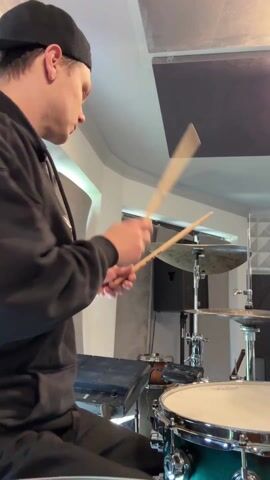 In a medium shot, a young man is drumming. He's wearing a black hoodie and a backwards baseball cap. He is fair skinned. He's playing a green snare drum, with a black electronic drum pad to his left, and a pair of cymbals in front of him. A black speaker and a stand with another cymbal are in the background. Above him are acoustic sound dampeners. The room is off white. He is actively playing the snare and cymbals with drumsticks.