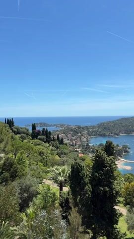 The video opens with a high-angle panoramic view of a lush green hillside sloping down towards a coastal town and a bay. The foreground is dominated by various trees and shrubs, including palm trees and tall cypress-like trees. In the mid-ground, a town with terracotta roofs is visible, nestled along the coastline. A sandy beach area and a calm, turquoise bay filled with numerous anchored boats and yachts are prominent features. Beyond the town and bay, a large, tree-covered peninsula or island extends into the deep blue sea. The sky is clear and bright blue with a faint contrail visible. The camera slowly pans to the right, gradually revealing more of the scenic coastal landscape.