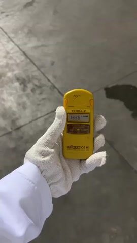 A gloved hand holds a yellow radiation detector. The detector displays the number 13.36. The background is a grey concrete floor. The video pans away from the detector to show a wider view of the concrete floor, which appears to be in an industrial setting with some structures visible in the distance. The floor has a few water stains.