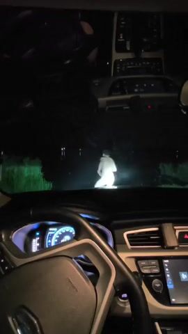 The video shows a view from inside a car at night. The dashboard and steering wheel are visible in the foreground. The headlights of the car illuminate a road ahead, where a person wearing a bright white outfit is standing. The person appears to be illuminated by the headlights, and as the car moves closer, the person moves and splashes water. The surrounding environment appears to be dark and possibly rural, with greenery visible on the sides of the road. The video is short and appears to capture a moment of encountering an unexpected figure on the road.