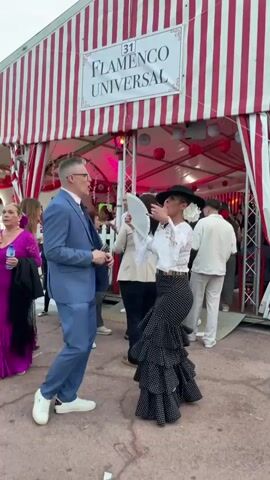 In front of a white and red striped tent labeled “Flamenco Universal,” a woman in a white blouse, polka-dotted layered skirt, and black hat dances with a white fan. She moves expressively with her arms raised. A man in a blue suit observes her, holding a glass. Other people mill about near the entrance of the tent.