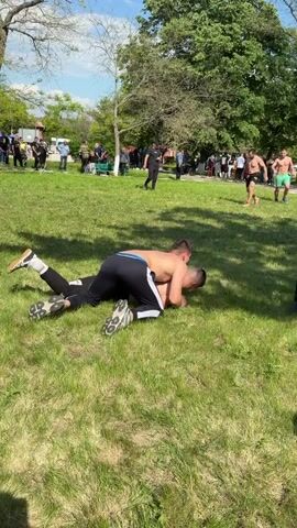 The video shows two men wrestling on a grassy field, part of a larger outdoor event with a crowd of people watching. The wrestlers are locked in a ground struggle, with one wearing black athletic pants with a white stripe and the other shirtless. In the background, other men are engaged in a similar wrestling match. Several spectators are standing around the perimeter of the field, watching the competitions. Trees provide a backdrop to the scene under a partially cloudy sky.