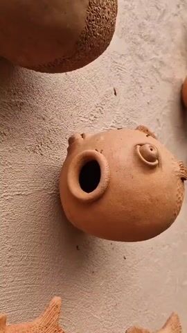 A close-up, vertical shot focuses on a collection of small, terracotta fish sculptures mounted on a textured, light-brown wall. The sculptures are all facing in various directions, some slightly angled towards the camera. They are uniformly brown and appear to be handmade, with visible texture and slight imperfections. Each fish has distinctive features: a round body, two small eye bumps, a fin on either side, and a textured area on its back, resembling scales or spiky skin. Most of the fish have an opening in the front, suggesting they might be planters or decorative holders. One prominent sculpture in the foreground, slightly to the left of the center, shows a wider opening and a more detailed eye. Another large sculpture is partially visible at the top left, also showing textured detailing. The background wall is uneven and has some small debris scattered on it, possibly dirt or natural material. To the right of the main cluster of fish, more sculptures are visible, receding into the distance and appearing smaller. Some greenery and a portion of another terracotta object are visible at the bottom right edge of the frame. The lighting is somewhat soft, with no harsh shadows.