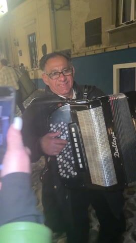 A man with short dark hair and glasses is seen from the chest up, playing an accordion on a cobblestone street at night. The accordion, labeled "Dallape," is black and silver, with visible red pleats. The man smiles as he plays. In the background, buildings with lit windows and painted blue walls are visible. Toward the end of the video, a hand holding a cell phone appears on the left, and someone hands the man a red rose.