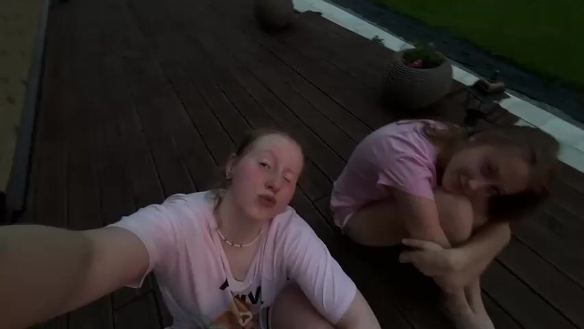The media file shows two young girls sitting on a wooden deck at dusk. The girl in the foreground is taking a selfie, making a playful face. She is wearing a pink t-shirt and a beaded necklace. The other girl is to her right, hugging her knees and looking down, appearing more introspective. She is also wearing a pink t-shirt and white shorts.

The setting is outdoors, with a wooden deck and some potted plants visible. In the background, there is a lawn and a fence, suggesting a residential area. The sky is cloudy and transitioning to twilight, with a hint of sunset colors peeking through in some frames. The overall mood is relaxed and casual, likely a moment captured during a summer evening. The camera movement is erratic in some shots, suggesting it might be a collection of clips from a phone camera, possibly filmed by one of the girls. There is no visible text in the media file. The location context is Gubakha, Russia, which is consistent with the outdoor setting and typical evening light for that region.