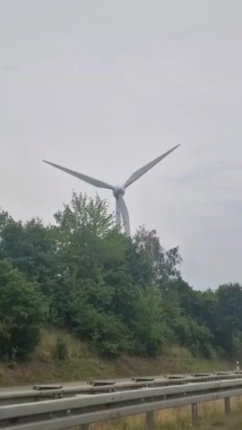 A wind turbine is visible against a cloudy sky, situated behind a line of trees. A road with a metal guardrail is in the foreground. A truck with a partially visible tank trailer drives past the wind turbine. Another wind turbine is visible in the distance to the right of the main turbine.