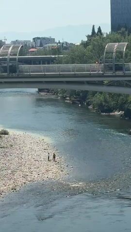 A wide river flows through a city. A pedestrian bridge spans the river. People are visible on the bridge and on the rocky shore of the river. A tall building stands on the far side of the river, behind trees. The sky is clear and blue.