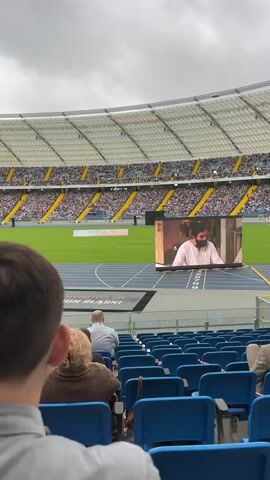 The video shows a wide shot of a stadium with a large crowd in attendance. In the foreground, the backs of several spectators are visible, seated in blue stadium chairs. Beyond them, a green athletic field is visible, with a large screen displaying a video. The screen shows a man in traditional clothing, then a scene of people working with fishing nets, and finally a close-up of what appears to be dead fish on the ground. The stadium seating extends up and around the field, and the sky above is overcast. There is a banner on the field with text in what appears to be Cyrillic script.