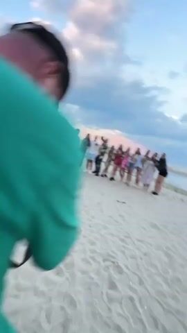 A group of approximately 15 people are standing in a line on a sandy beach posing for a photo. The person taking the photo is a man wearing a green shirt and black baseball cap. He holds a camera in his hands and is focused on the group. The group of people are dressed in casual clothing, and they are smiling and pointing towards the camera. The sky is partly cloudy with shades of pink and blue, suggesting it may be sunset. In the background, there is a body of water and some sand dunes. The scene suggests a fun and celebratory atmosphere, possibly a group of friends or colleagues enjoying a gathering at the beach.