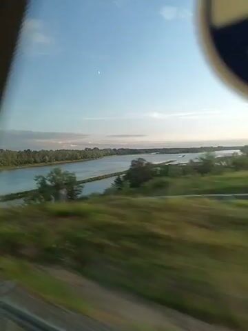 A wide shot captures a river under a bright, partly cloudy sky. The water is a deep blue, reflecting the sky. A long, narrow strip of land covered in grass cuts across the river, dividing it into two channels. Trees line the far bank of the river. The foreground shows a blurred section of what appears to be a train or vehicle, suggesting movement. The overall scene is serene and natural, bathed in the soft light of what seems to be either sunrise or sunset.