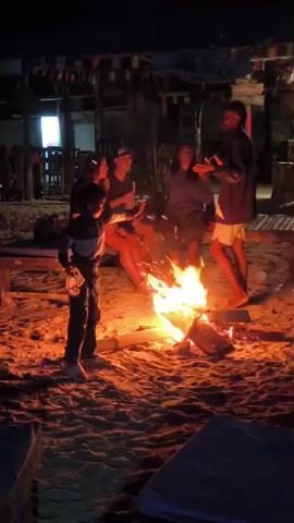 The video depicts a nighttime beach scene with a group of people gathered around a campfire. Four individuals are seated on a low wooden bench, while another person stands beside the fire, gesturing with their hands. A child is also visible, moving around in the foreground near the fire. The setting appears to be a beachside area with a wooden structure, possibly a bar or restaurant, visible in the background, adorned with strung flags. The scene is illuminated by the glow of the fire, casting long shadows on the sandy beach.
